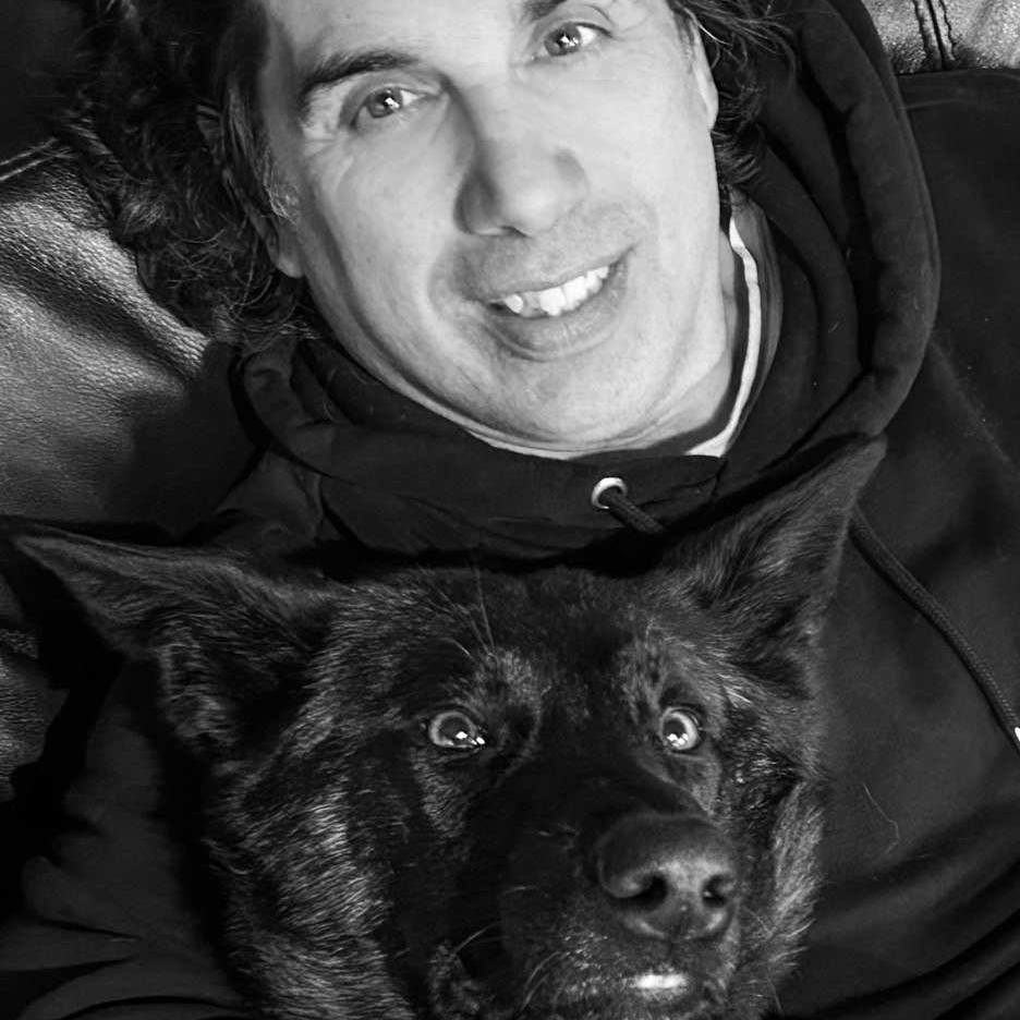 Black-and-white portrait of a man smiling with his black dog, Leo, both lying down and looking into the camera.