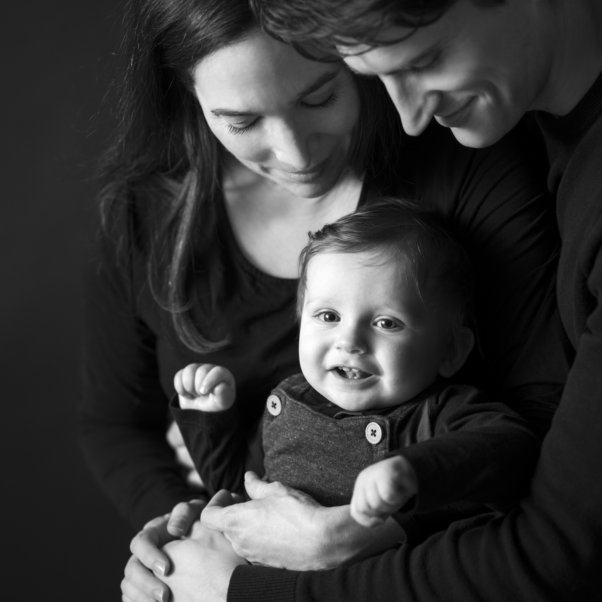 Family with Baby – Black-and-White Fine Art Portrait Black-and-white portrait of a smiling baby held in the arms of loving parents, all wearing dark clothing, by Jerome Art Photography.