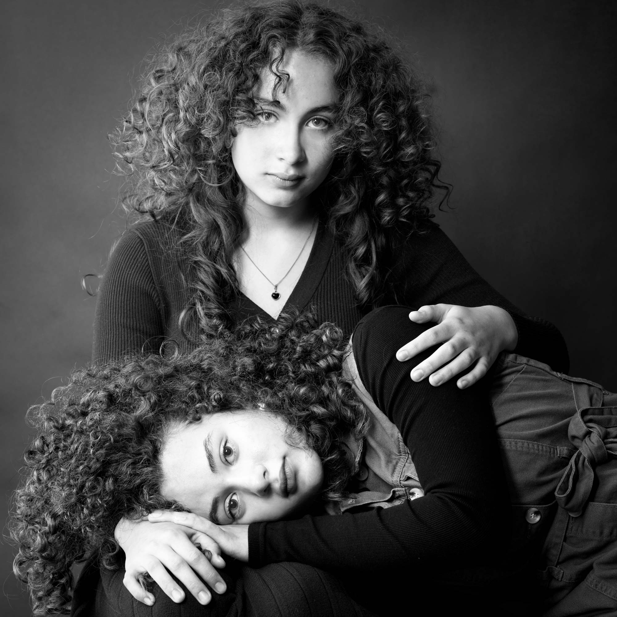 Black-and-white portrait of two young girls with curly hair, one sitting and the other resting her head on her sister’s lap, by Jerome Art Photography.